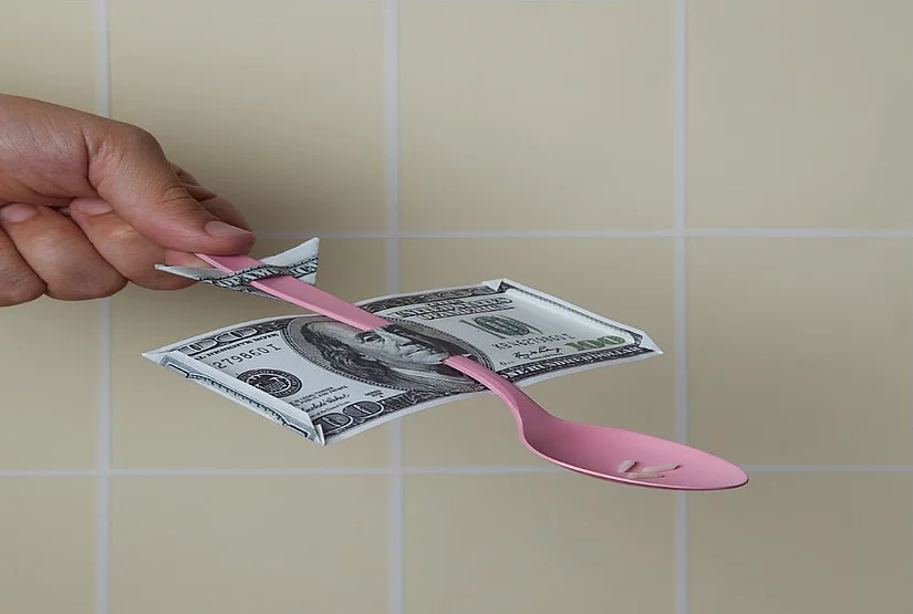 Una mano sujeta una cuchara de plástico que contiene tres granos de arroz, mientras el mango de la cuchara atraviesa a un billete de 100 dólares.